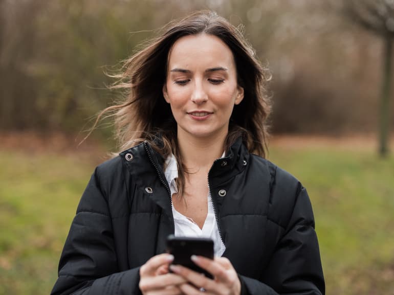 Mørkhåret kvinde kigger interesseret på Falck Sundhedshjælp - Online-læge på sin telefon.