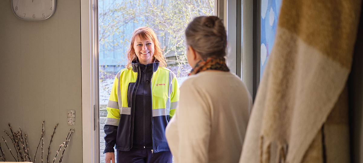 En Falck-medarbejder i gul refleksjakke står smilende i en åben dør og bliver mødt af en ældre kvinde indenfor i hjemmet.