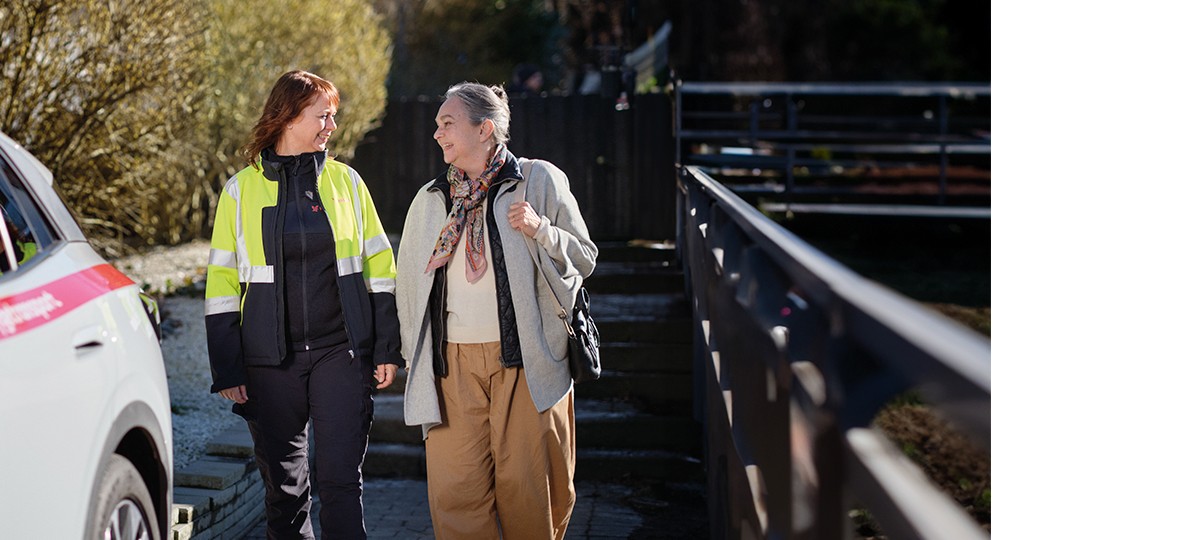 En falck medarbejder i gul refleksjakke står smilende sammen med en ældre kvinde foran en sygetransportbil. 